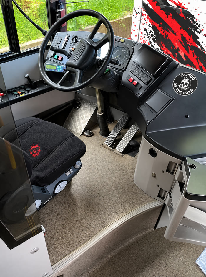 Fahrerplatz des Tattoo-Busses: schwarzer Sitzbezug mit rotem Totenkopf, Tattoo-On-The-Road-Aufkleber am Armaturenbrett.