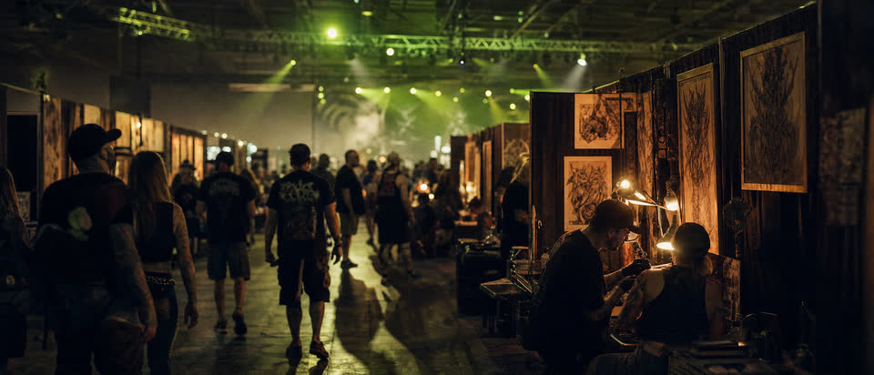 Blick in eine Tattoo-Convention-Halle, Reihen von Ständen unter grünem Traversenlicht.